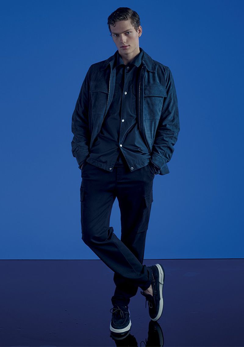 A young adult male model poses in a dark blue shirt and pants with a blue jacket, set against a matching blue backdrop.