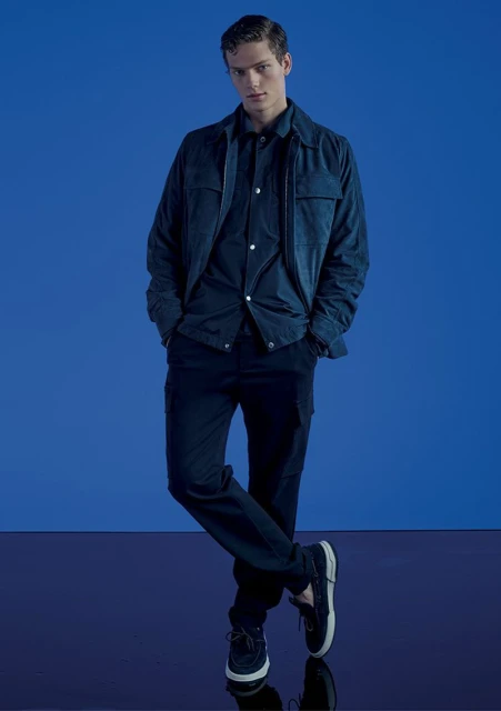A young adult male model poses in a dark blue shirt and pants with a blue jacket, set against a matching blue backdrop.