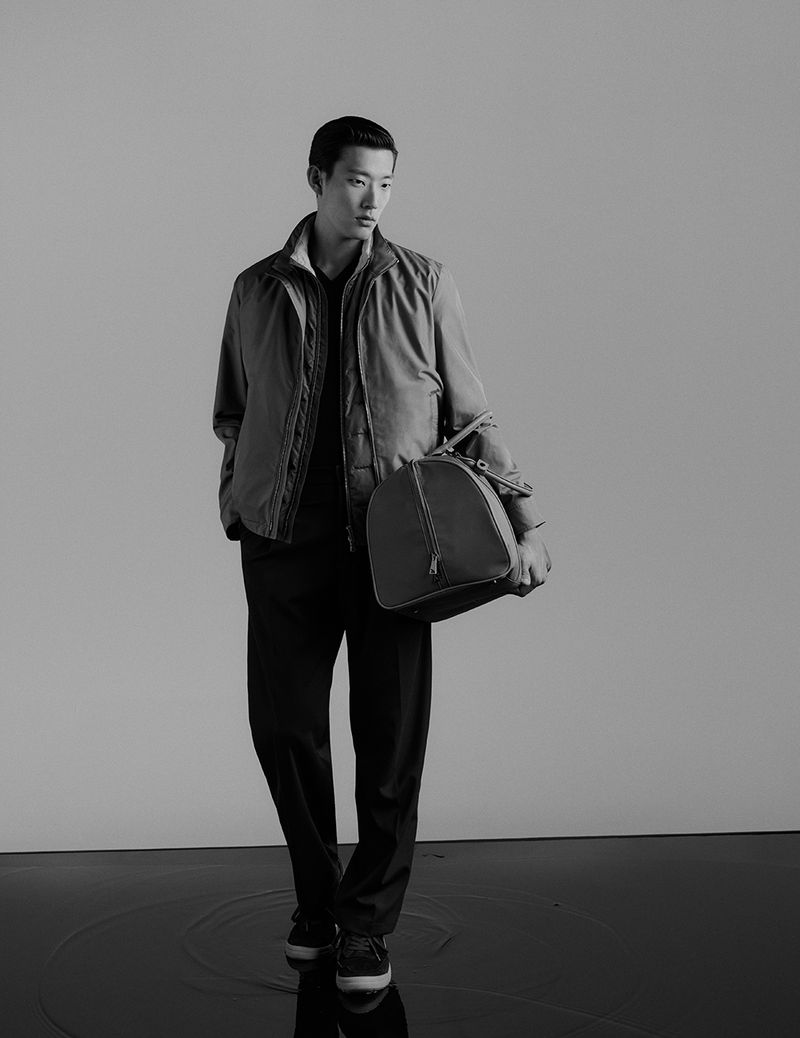 Monochrome portrait of an adult male in a jacket and slacks holding a duffel bag in a brightly lit indoor photo studio.