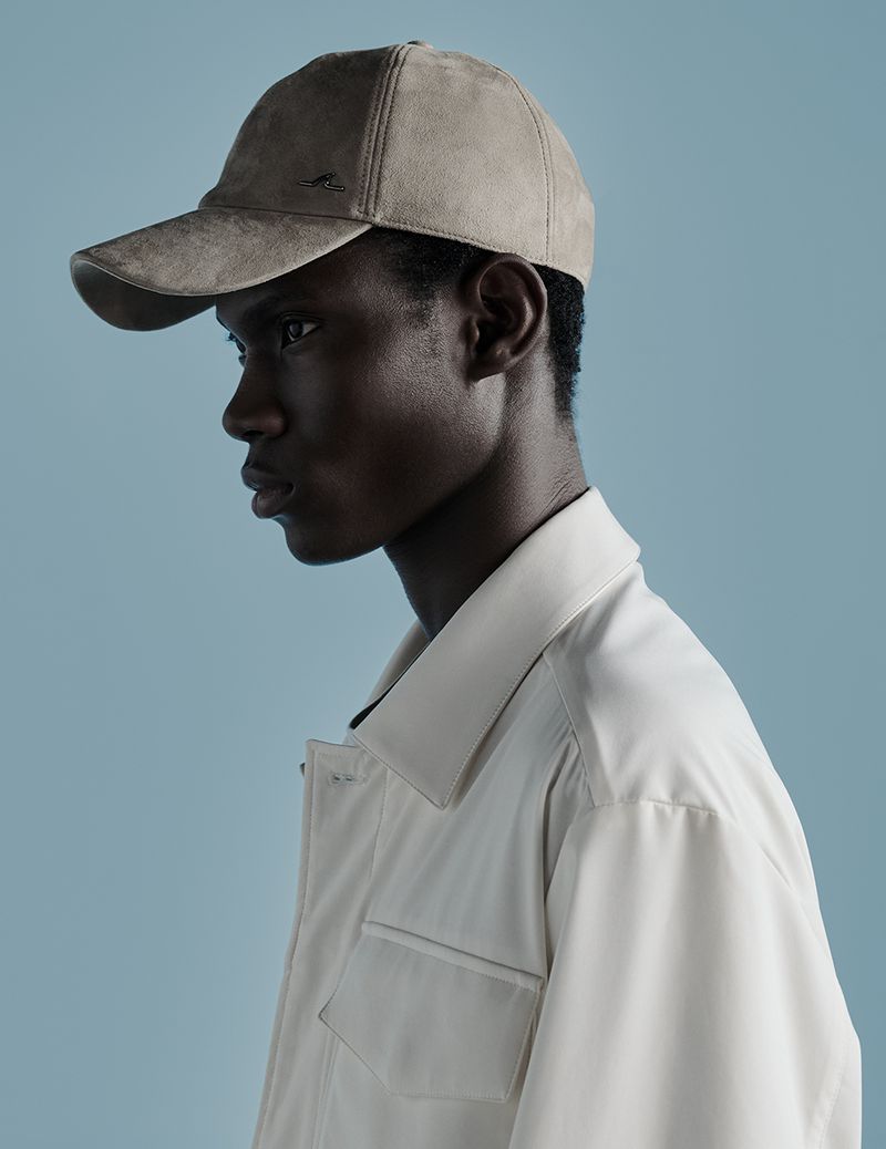 Profile of a young adult male wearing a tan baseball cap and an off-white jacket against a light blue background in a studio setting.