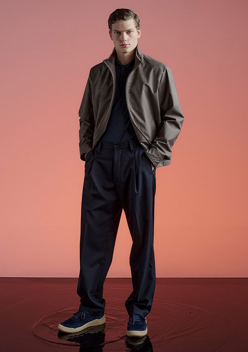 Full-length shot of an adult male fashion model in a taupe jacket, navy pants, and navy shoes in a photo studio against a peach-colored background.