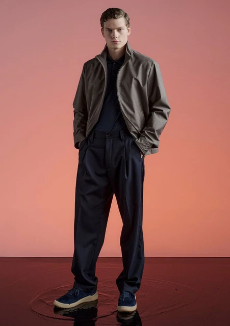 Full-length shot of an adult male fashion model in a taupe jacket, navy pants, and navy shoes in a photo studio against a peach-colored background.