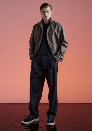 Full-length shot of an adult male fashion model in a taupe jacket, navy pants, and navy shoes in a photo studio against a peach-colored background.