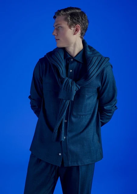 Studio shot of a young adult male wearing a blue collared shirt and pants, a matching sweater draped over his shoulders against blue backdrop.