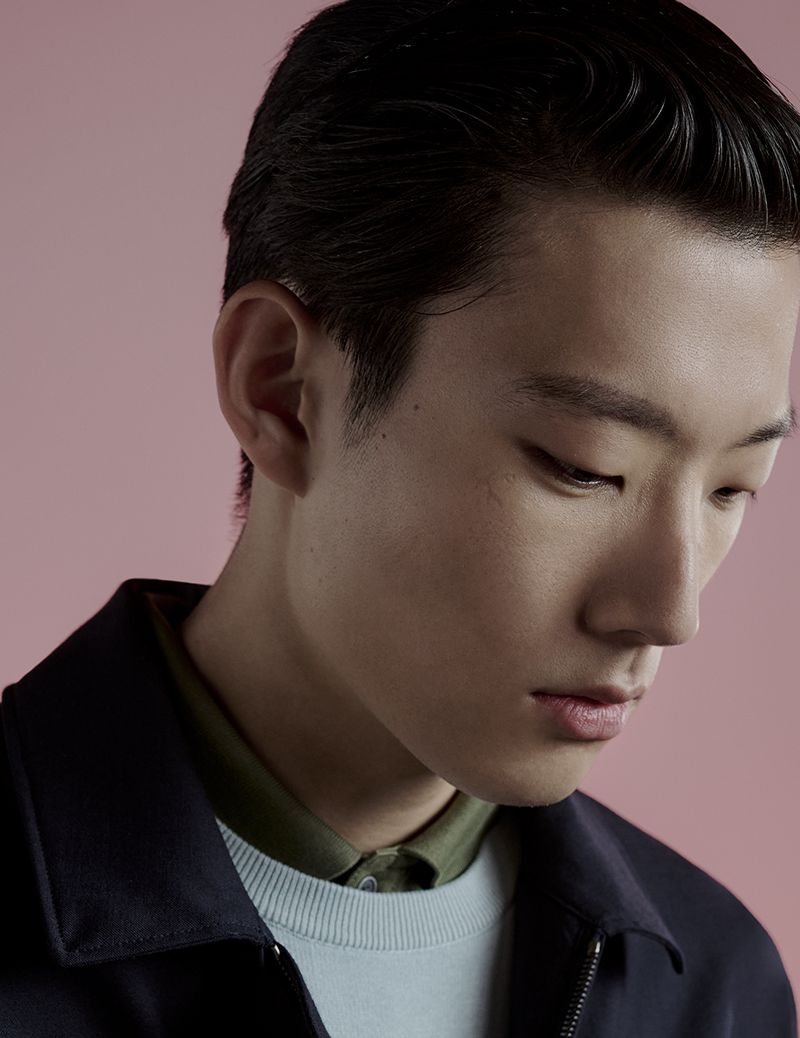 Close-up studio portrait of an Asian adult male model with dark hair, wearing a navy jacket over a light blue sweater. Soft pink background.