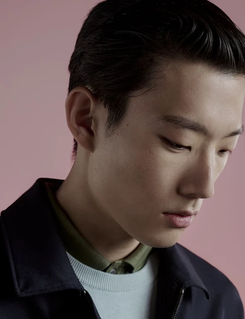 Close-up studio portrait of an Asian adult male model with dark hair, wearing a navy jacket over a light blue sweater. Soft pink background.