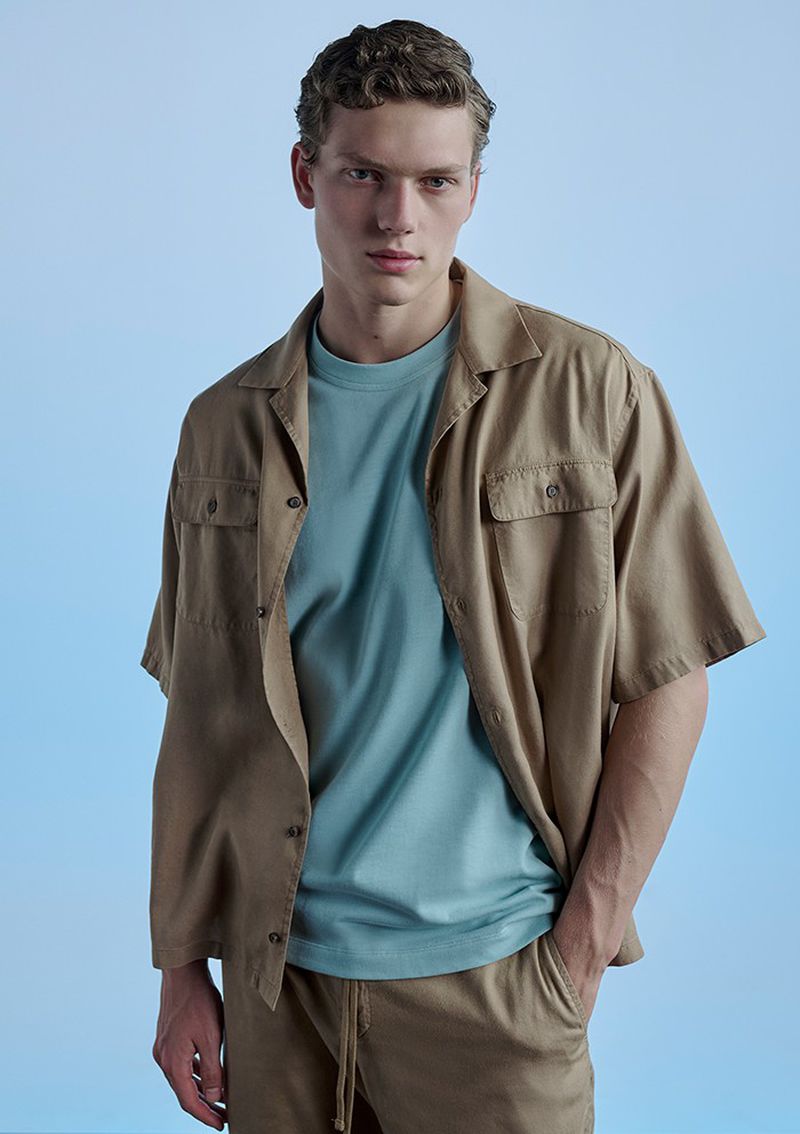 A young adult man poses in a tan camp shirt, light blue t-shirt, and tan shorts against a light blue backdrop in a studio setting.