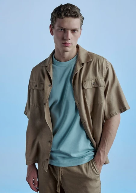 A young adult man poses in a tan camp shirt, light blue t-shirt, and tan shorts against a light blue backdrop in a studio setting.