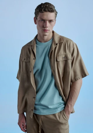 A young adult man poses in a tan camp shirt, light blue t-shirt, and tan shorts against a light blue backdrop in a studio setting.
