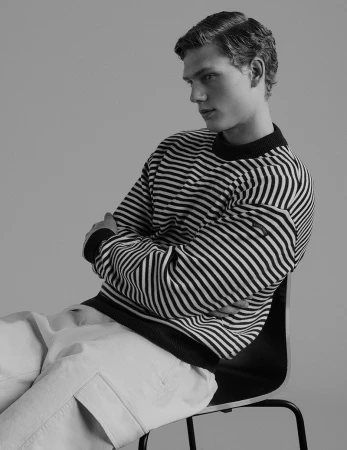 A young male model seated on a dark chair wearing a striped knit sweater against a grey studio backdrop.