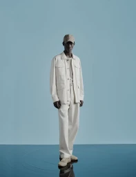Model standing on a glossy floor against a blue background wearing an off-white jacket, trousers, cap, and sneakers.