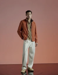 Full-length portrait of a male model wearing an orange jacket, tan zip-up top, and white trousers against a pink studio backdrop.