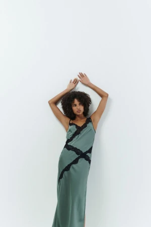 A woman with curly hair leans against a white wall, wearing a long, light-green dress with black lace details and black straps, indoor setting.