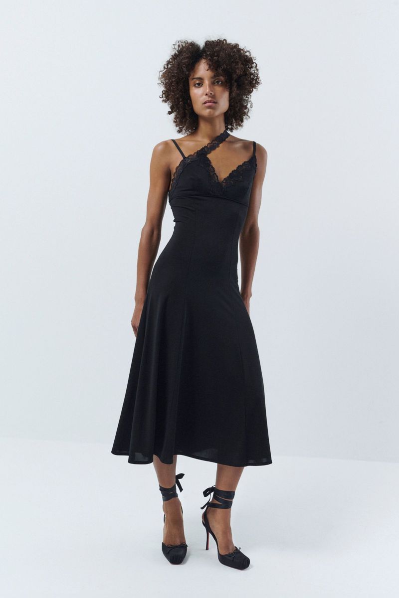 A slender African American woman poses in a black lace-trimmed dress and matching heels, against a stark white backdrop in a studio shot.
