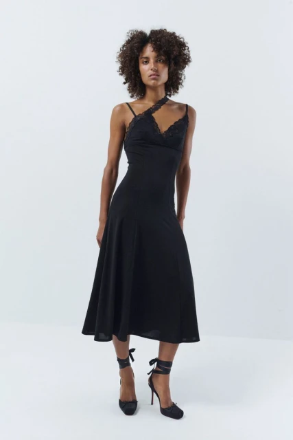 A slender African American woman poses in a black lace-trimmed dress and matching heels, against a stark white backdrop in a studio shot.