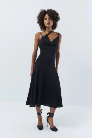 A slender African American woman poses in a black lace-trimmed dress and matching heels, against a stark white backdrop in a studio shot.
