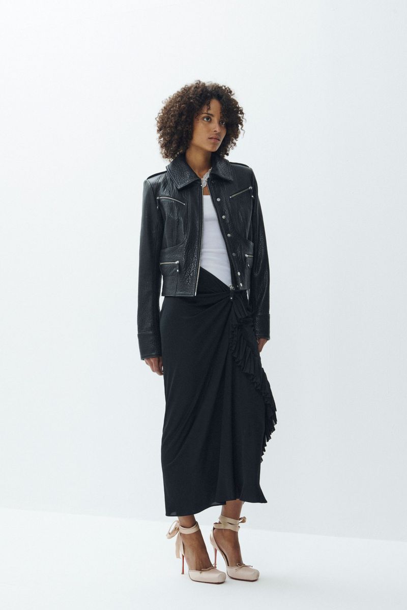 Model with curly hair wearing a black leather jacket and black skirt against a white studio background.