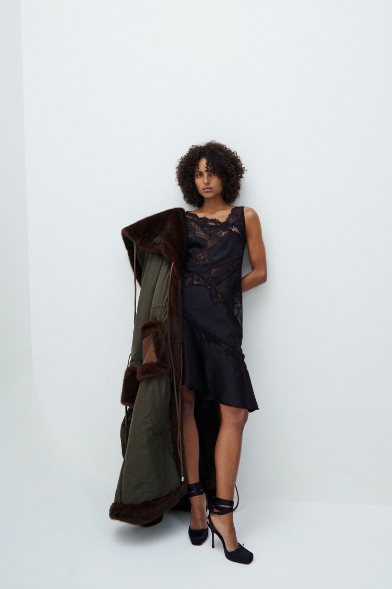 Elegant woman with curly hair poses indoors wearing a black lace dress and heels, holding an olive green fur-trimmed coat.