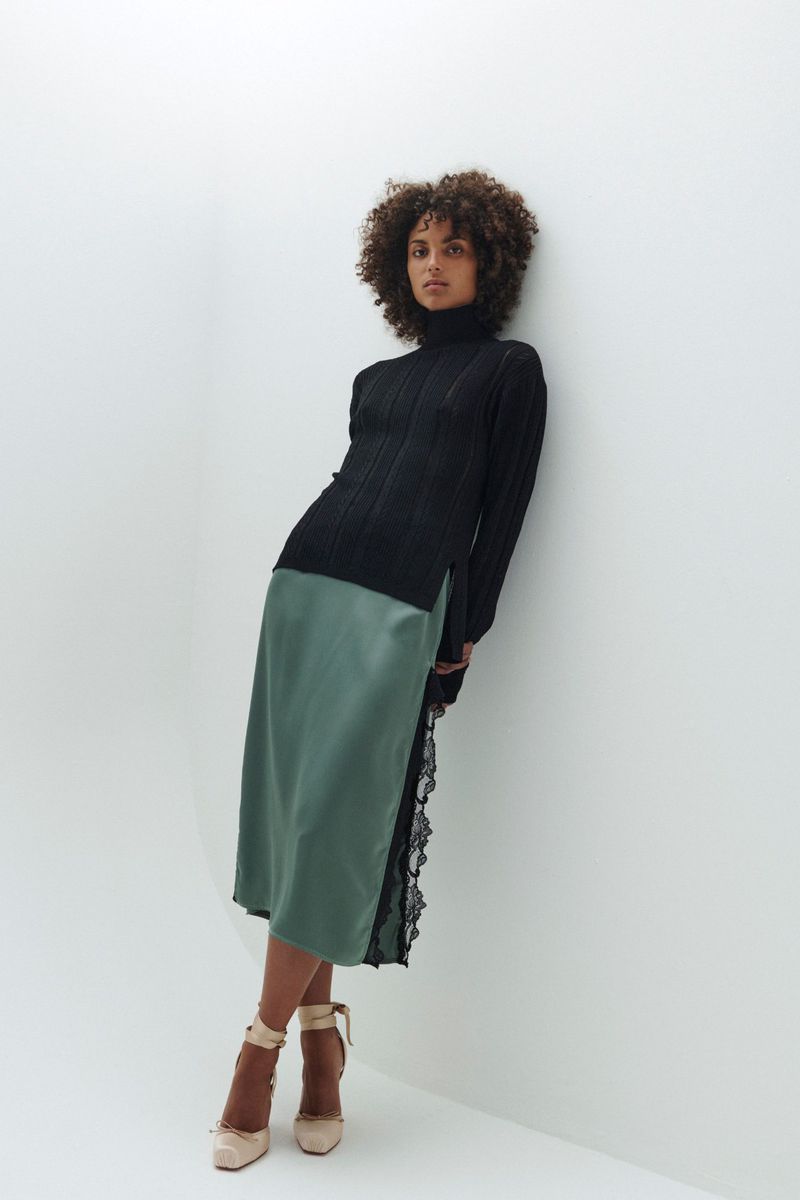 A woman with curly hair poses against a white wall wearing a black turtleneck sweater and a satin, lace-trimmed skirt with high heels.