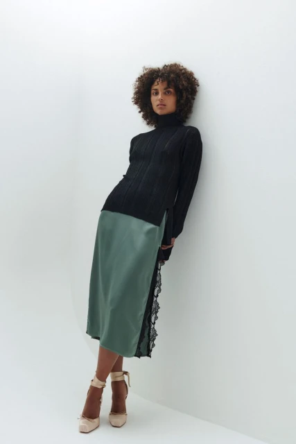 A woman with curly hair poses against a white wall wearing a black turtleneck sweater and a satin, lace-trimmed skirt with high heels.