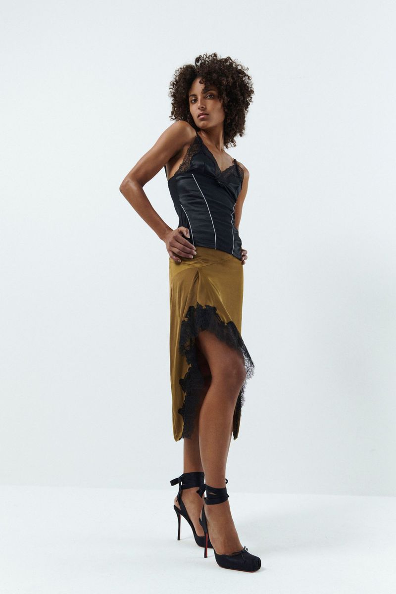Model in a black satin top and olive midi skirt standing against a white seamless background for Vaillant Pre-Fall 25.