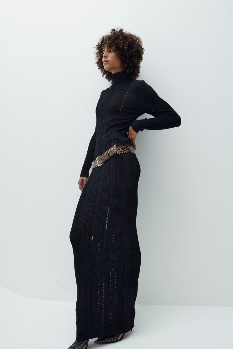 A woman in a black, long-sleeved maxi dress and patterned belt poses in front of a white wall in a fashion photography studio.