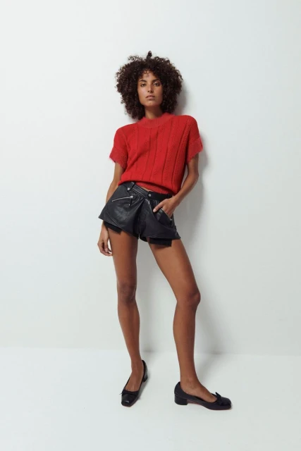 Fashion model poses in studio wearing a red knit sweater, black leather shorts, and black flats against a white backdrop.