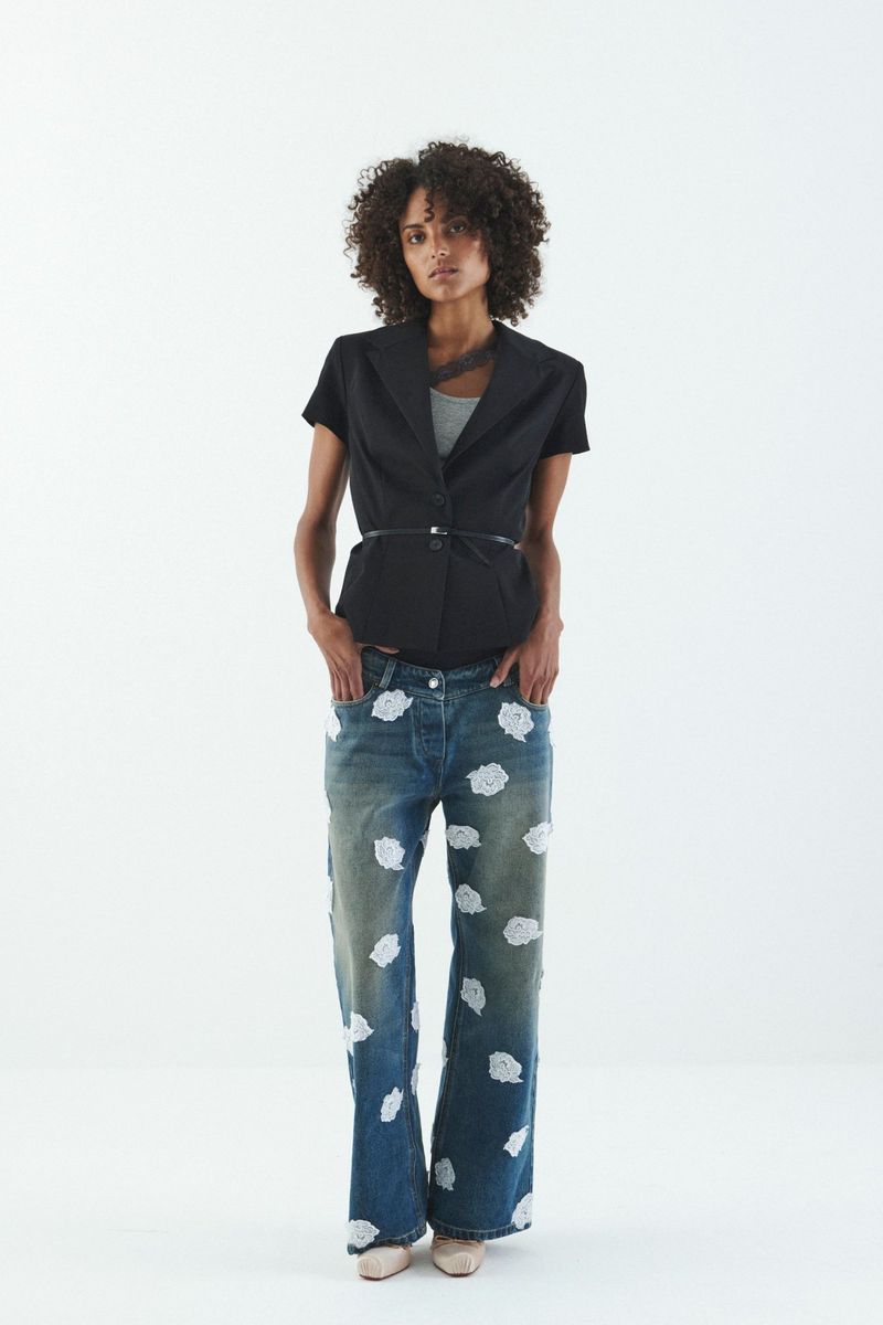 A woman with curly hair is posing in a short-sleeved black blazer, gray tank top, and blue jeans with cloud-like floral designs against a white backdrop.