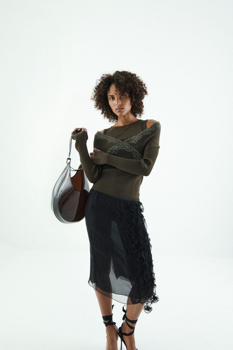 A curly-haired woman in a fashionable olive top and black skirt holds a large leather bag, posing against a clean, white backdrop.
