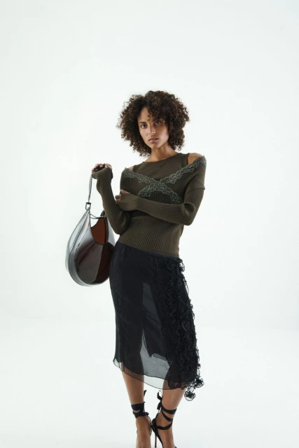 A curly-haired woman in a fashionable olive top and black skirt holds a large leather bag, posing against a clean, white backdrop.