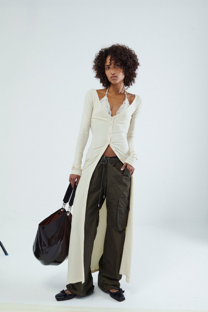A woman poses in a long cardigan, cargo pants, and black tote bag against a white backdrop in a fashion editorial-style full shot.