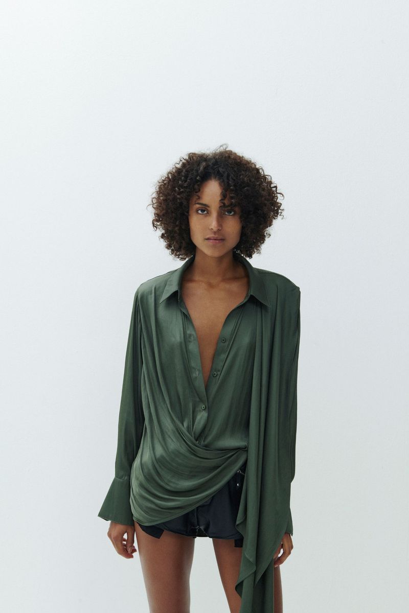 Adult female with curly hair poses in an olive-green draped button-down shirt with black shorts against a white backdrop in a studio.