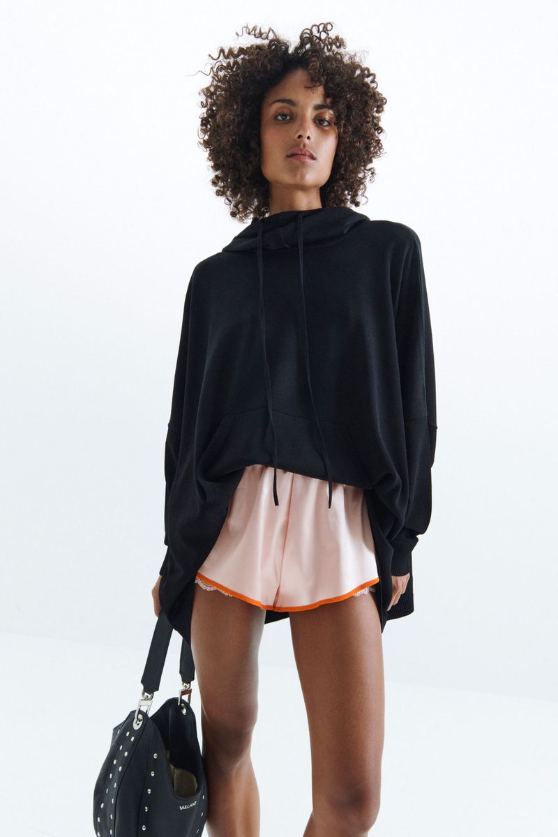 Adult female with curly hair posing in a black hoodie and peach shorts, holding a black studded bag against a white backdrop.