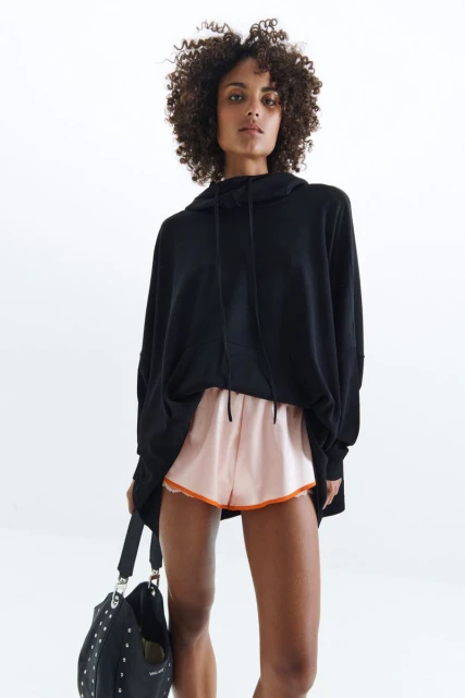 Adult female with curly hair posing in a black hoodie and peach shorts, holding a black studded bag against a white backdrop.