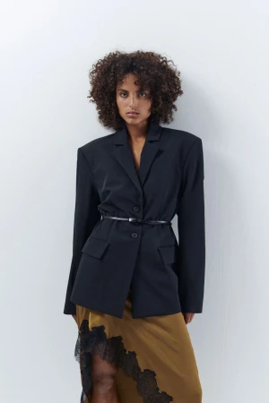 Model with dark curly hair wearing a structured black blazer and brown lace skirt against a white wall.