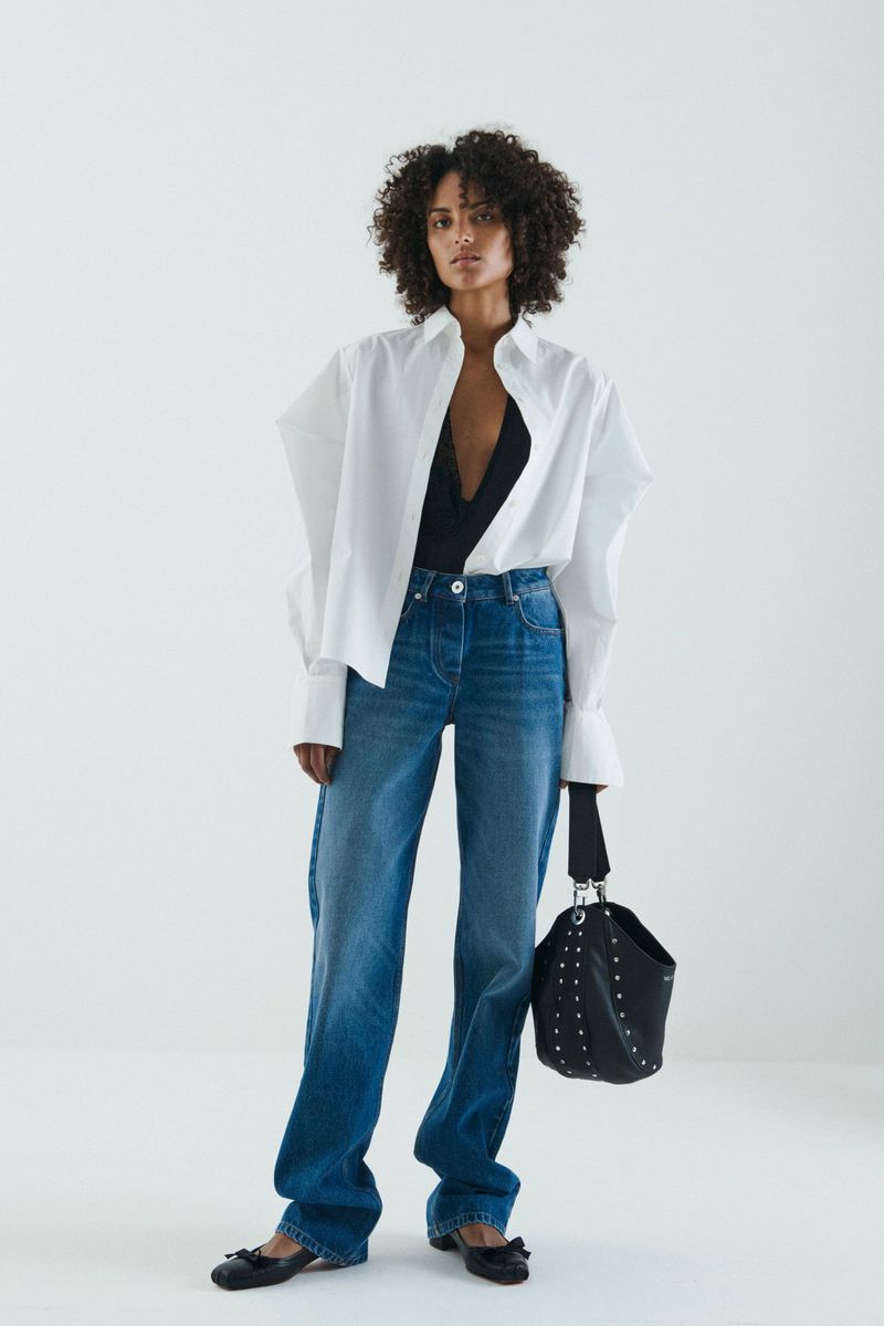 Woman in a black top, white shirt, and blue jeans stands in a white studio holding a studded black bucket bag in a fashion shot.
