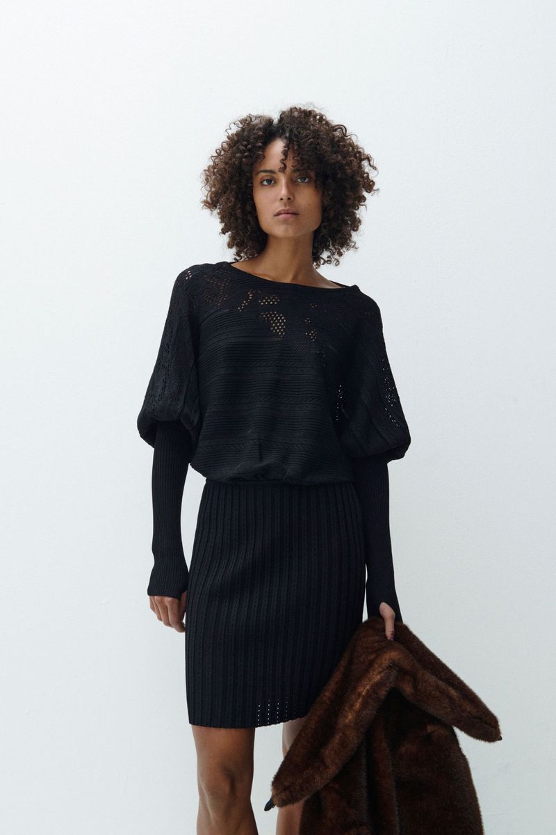 An adult female model poses in a black knit dress against a white background, holding a brown faux fur coat, in a fashion-forward shot.