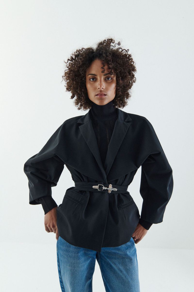 A curly-haired woman in a black blazer with a decorative belt, jeans, and a black turtleneck poses in a fashion studio with white walls.