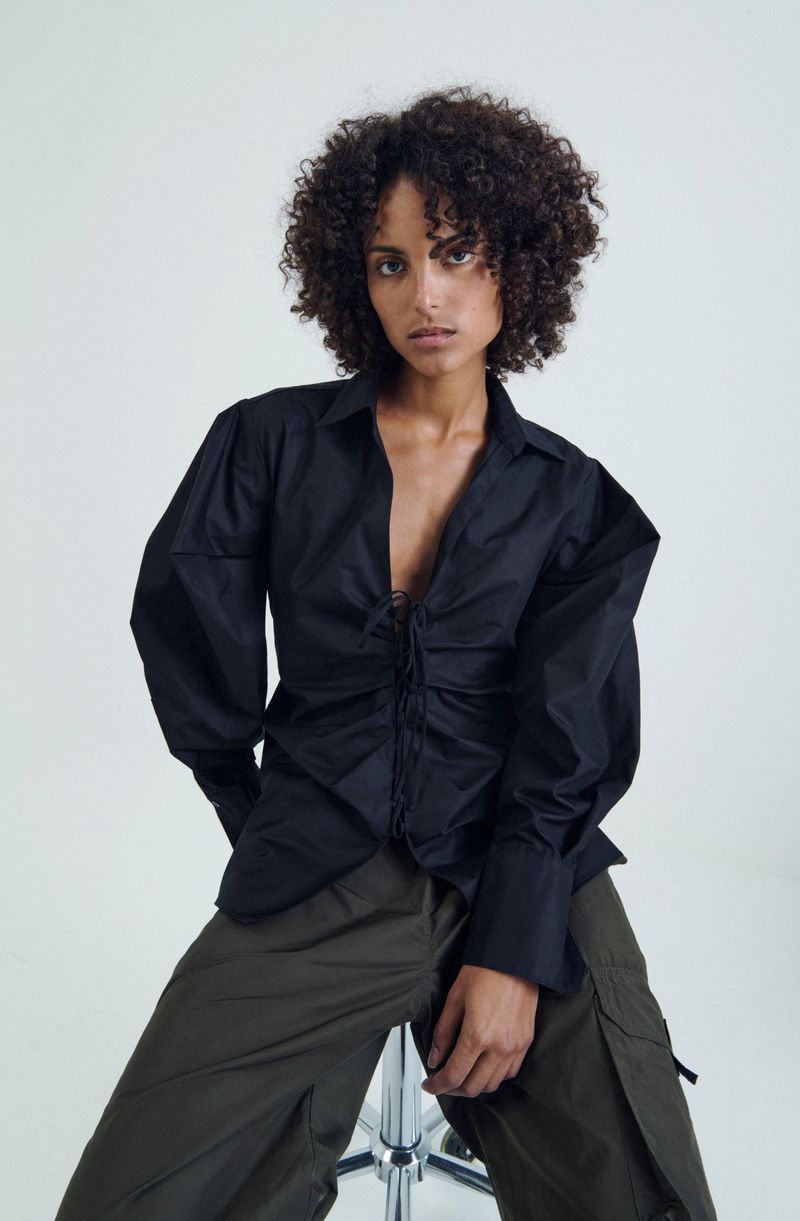 A woman with curly hair poses in a black blouse and green pants, sitting on a stool against a bright white backdrop in a fashion shot.