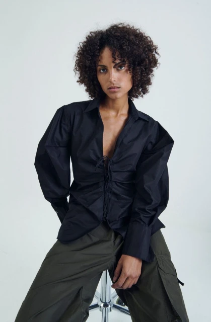 A woman with curly hair poses in a black blouse and green pants, sitting on a stool against a bright white backdrop in a fashion shot.
