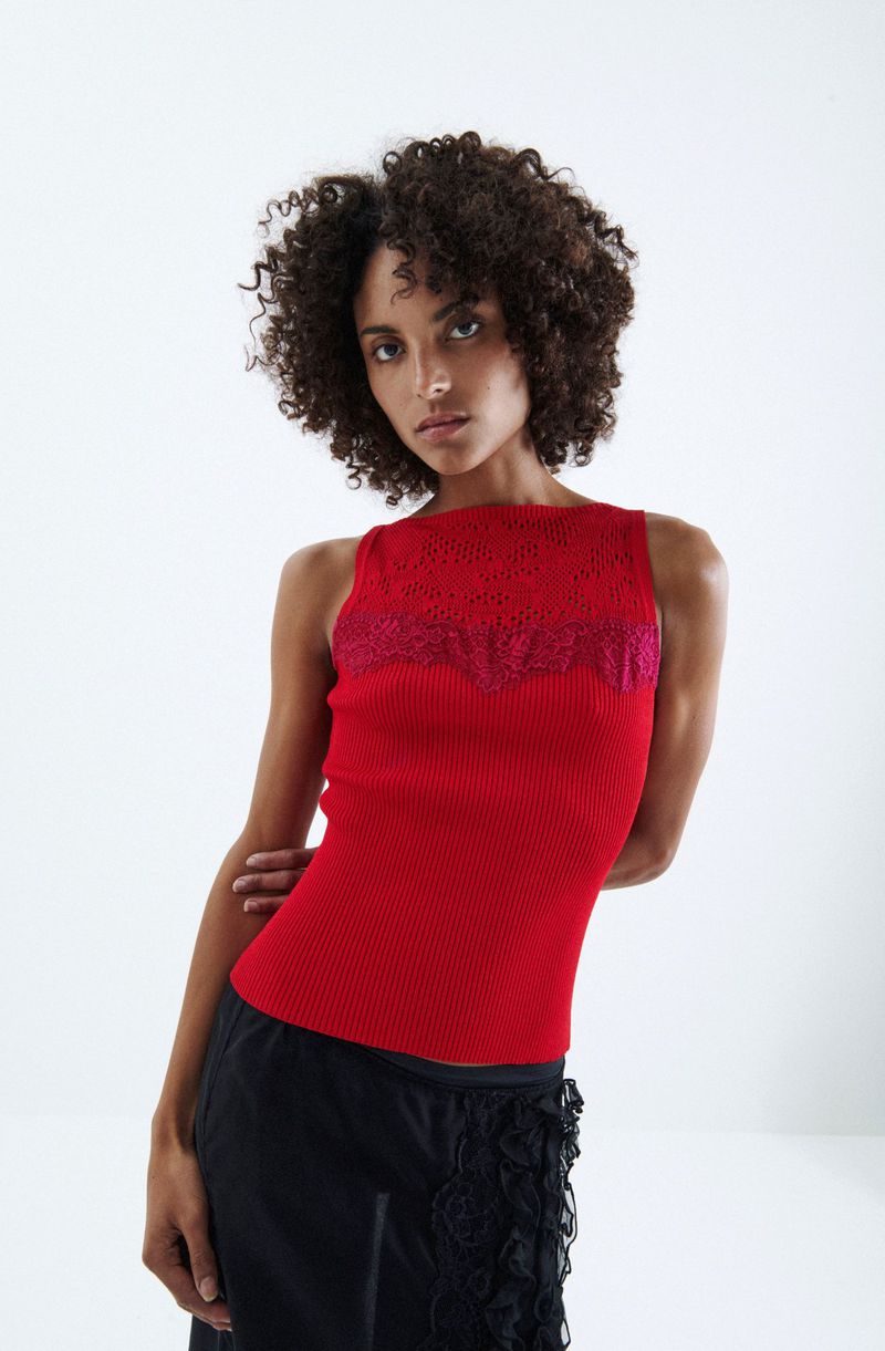 A brunette woman with curly hair poses in a red ribbed tank top with a lace detail at the neckline against a bright white background.