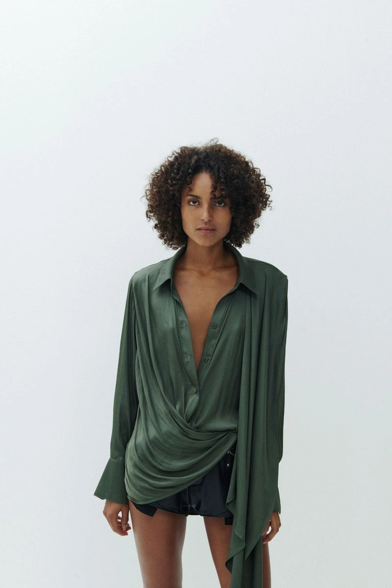Model with dark curly hair wearing an olive green silk shirt against a white seamless background