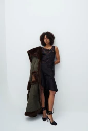 Model in black lace slip dress holding a long dark coat against a white background for Vaillant Pre-Fall 25.