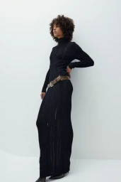 Model with curly dark hair wearing a black ribbed turtleneck dress and patterned belt against a white backdrop.
