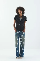Model with dark curly hair wearing a short-sleeved black blazer and patterned denim jeans against a white background