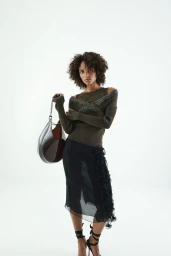 Model with curly hair wearing an olive green top and black skirt holding a large dark handbag against a white background