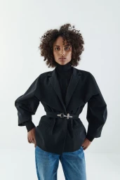 Model with curly dark hair wearing a black blazer and blue jeans against a plain white background