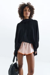 Model with curly hair wearing an oversized black hoodie and pink shorts holding a black studded handbag against a plain white background