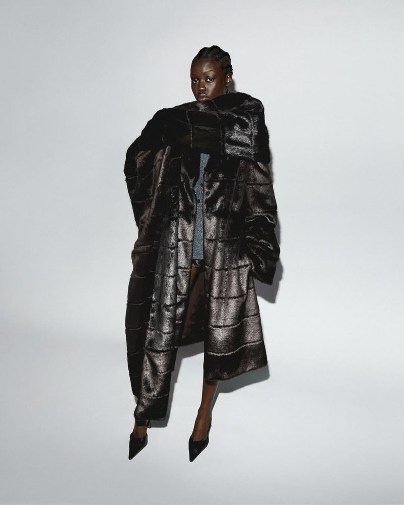 Fashionable Black woman poses in a dark patterned coat, black high heels against a white backdrop in a studio setting, showcasing elegant style.