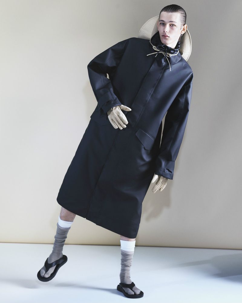 Fashion editorial shot of a slender male model in a black trench coat, socks, and sandals, with a straw hat in a studio setting.